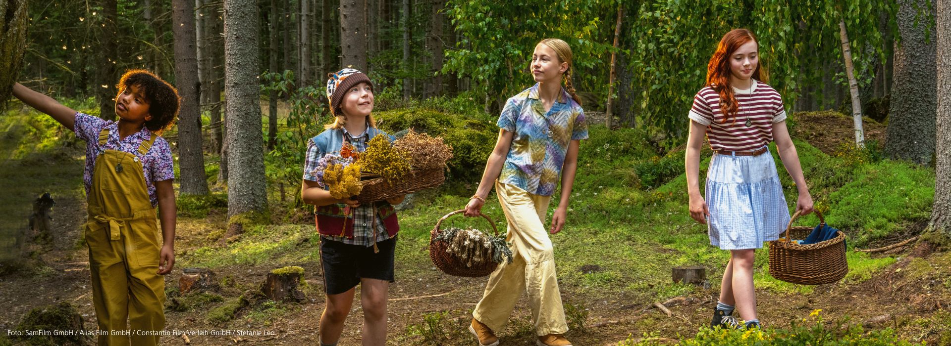 Szene aus dem Film ‘Ein Mädchen namens Willow’: Die Hexenfreundinnen Lotti, Gretchen, Valentina und Willow spazieren mit Körben voller Blumen durch einen sonnigen, magischen Wald.