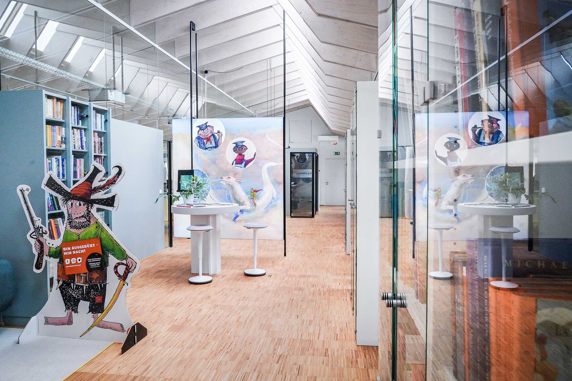 Foto des Verlags: Heller, moderner, freundlicher Flur mit Holzfußboden, bunten Bücherregalen, Glaswänden und Aufstellern von Kinderbuchfiguren.